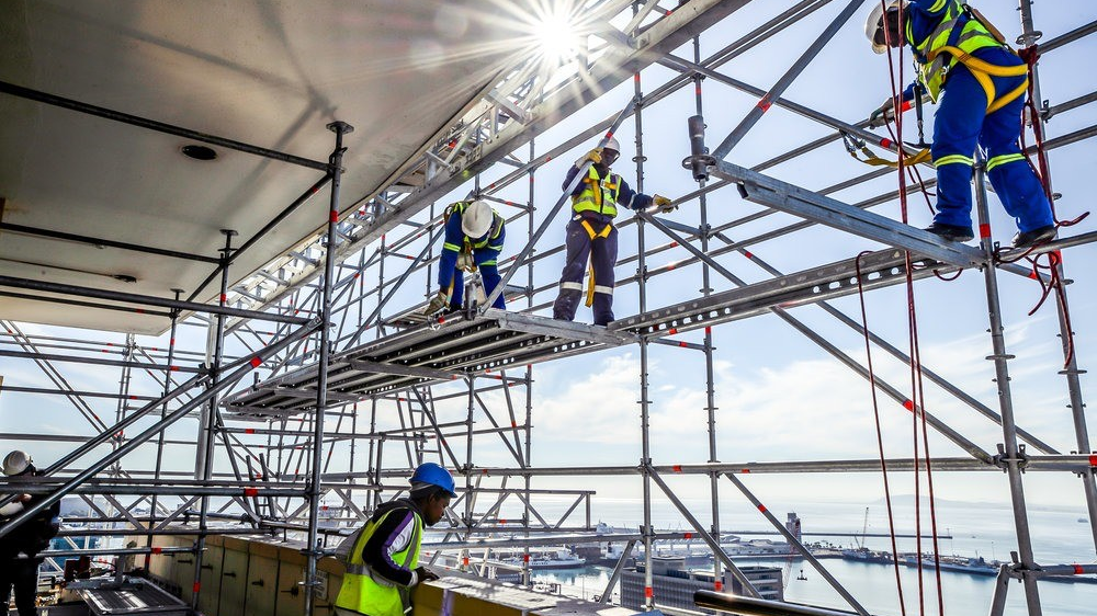 Certified scaffolders in full PPE and harnesses erecting access scaffolding above a harbour industrial site